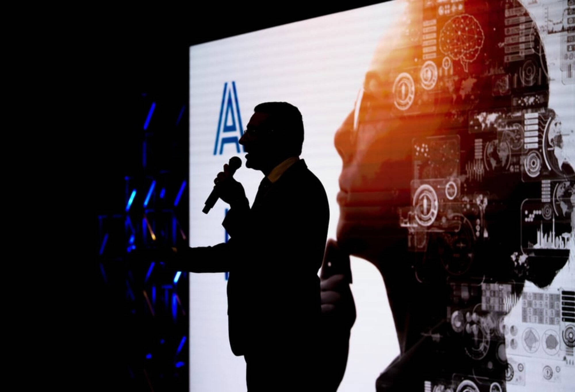 Dean Marouane Kessentini holding a microphone stands in silhouette against a large digital display featuring a human profile with technology and circuit graphics